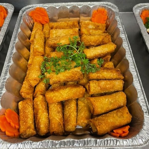 Golden-brown egg rolls with carved carrots and parsley in a foil catering tray.