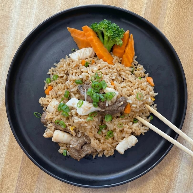 Beef and seafood fried rice with broccoli, carrots, and green onions on a black plate with chopsticks.