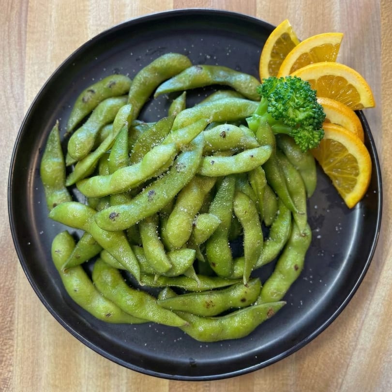 Salted edamame pods on a black plate, garnished with broccoli and orange slices.