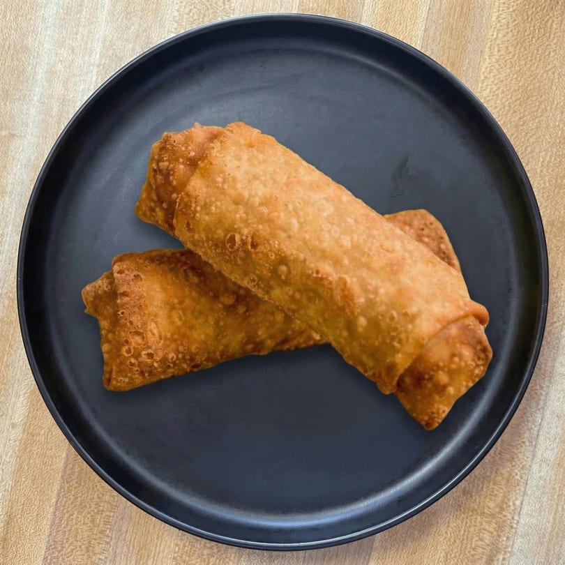 Two golden-brown egg rolls are served on a dark gray plate on a light wooden table.