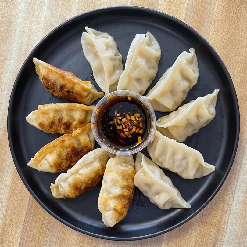 A black plate with a ring of steamed and pan-fried dumplings surrounding a cup of dipping sauce.