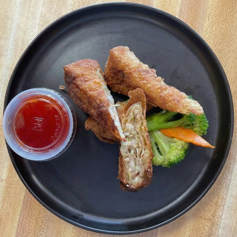 Crispy fried egg rolls with sweet chili sauce, broccoli, and carrots on a black plate.