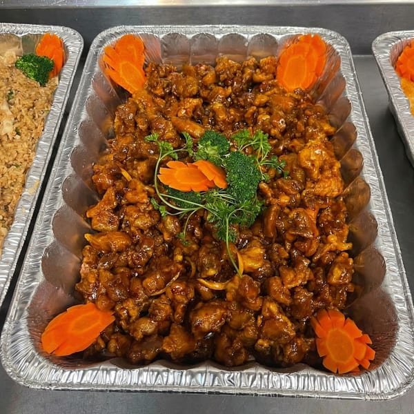 A foil tray of orange chicken with broccoli, parsley, and carved carrots, next to fried rice.